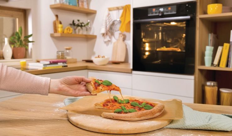 Žena si berie ksok čerstvo urobenej pizze, zatiaľ čo sa druh pripravuje v parnej rre.