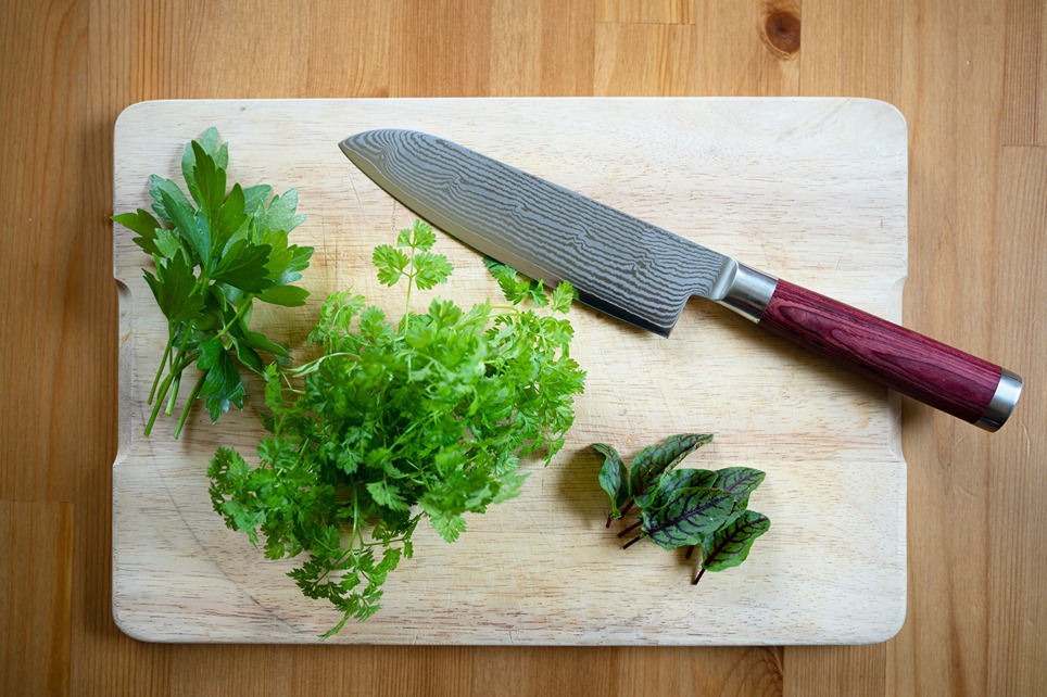 Kuchynský nôž z damaškovej ocele položený na doske na krájanie s bylinkami.